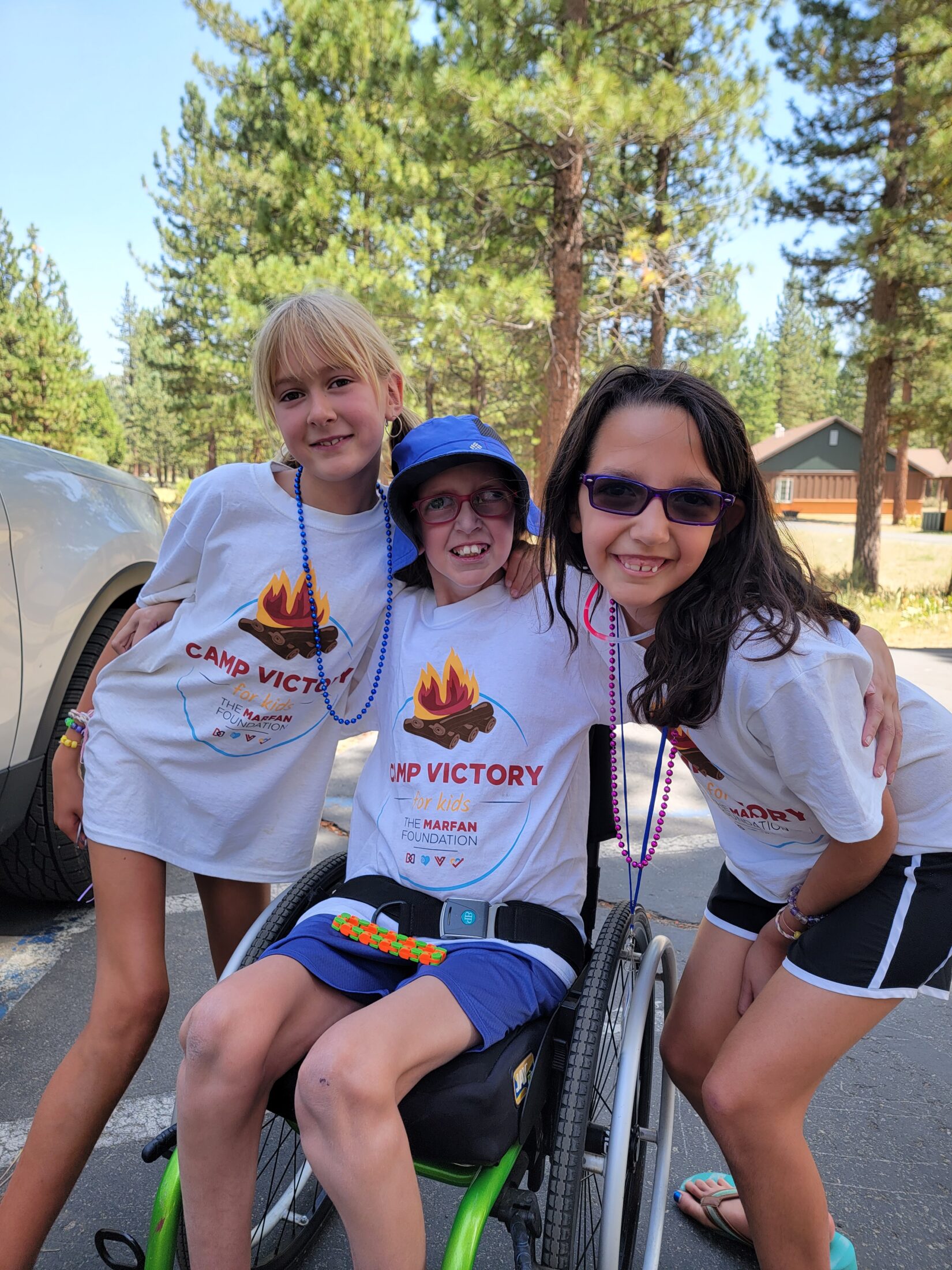 three kids, two standing, one in seated in wheelchair, huddled together and smiling while wearing matching camp victory t-shirts surrounded by trees