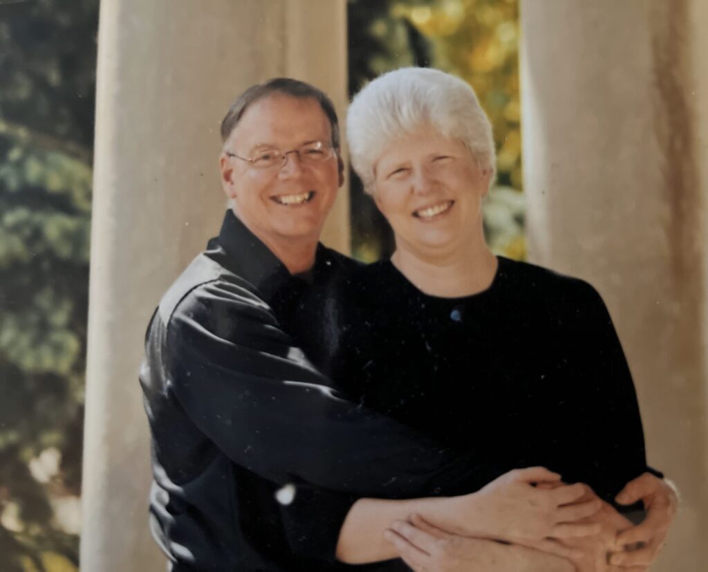 Donald Albrecht with arms around his wife Justice Adrienne Albrecht while both smiling in front of a nature scene. 