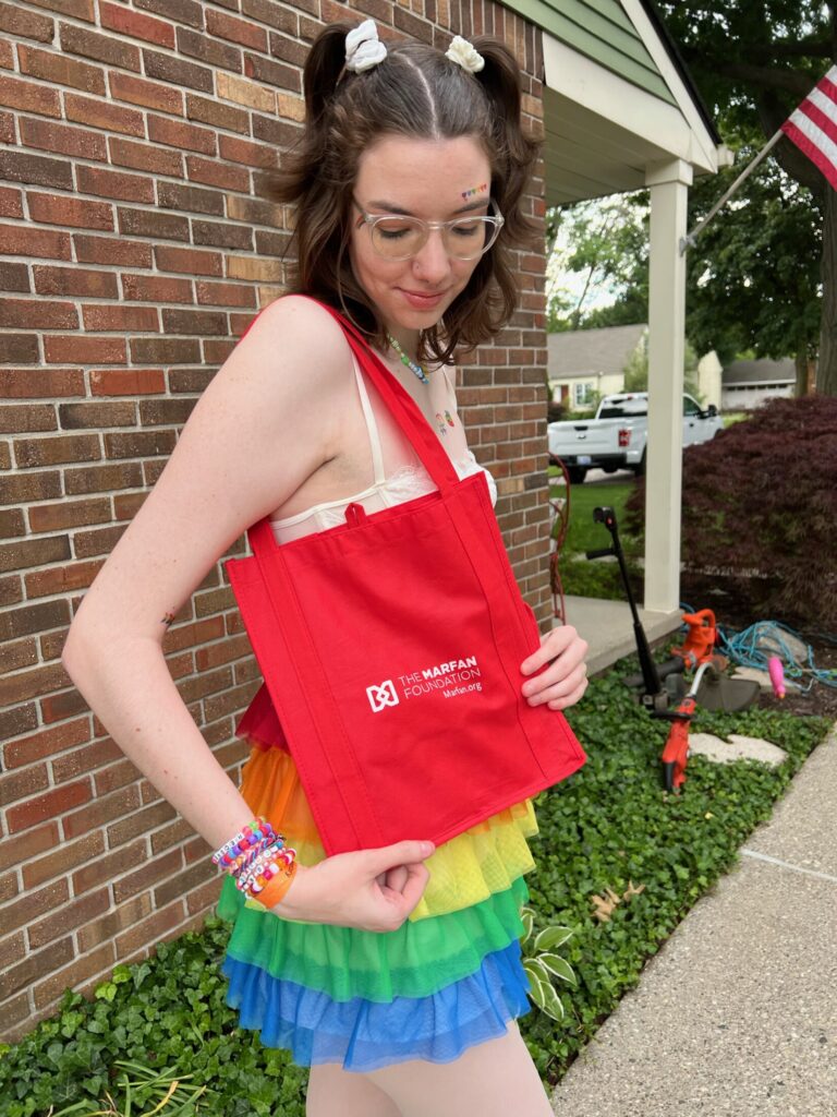 Grace Meyers dressed in Pride gear looks at a Marfan Foundation tote bag 