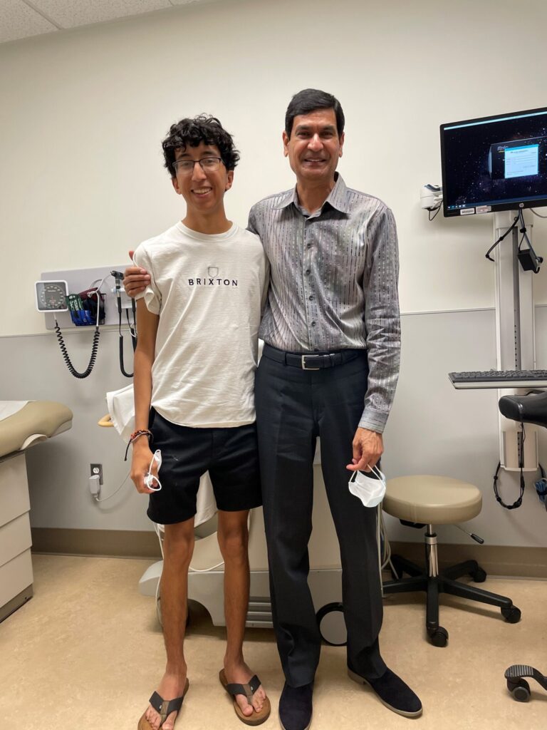 Young adult stands in hospital setting, with his pediatric cardiologist, both smiling 