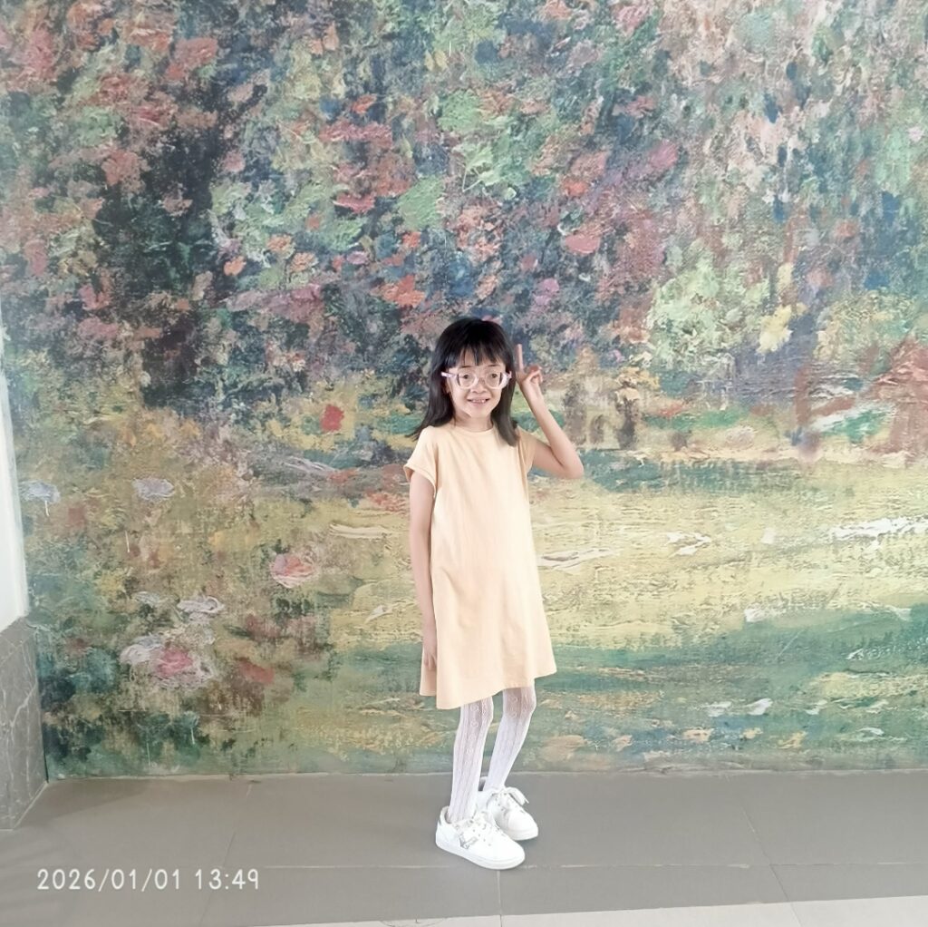 Destinee Jaye poses in a light yellow dress in front of a pastel background wearing glasses and flashing the peace sign. She appears to have some classic Marfan features such as long thin limbs. 