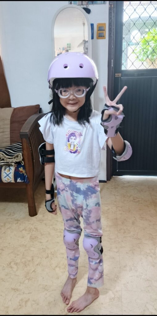 Destinee Jaye poses wearing a helmet and arm/wrist protectors for a sport in multicolor tie dye pants and white shirt, flashing the peace sign. Destinee lives with early-onset Marfan syndrome.