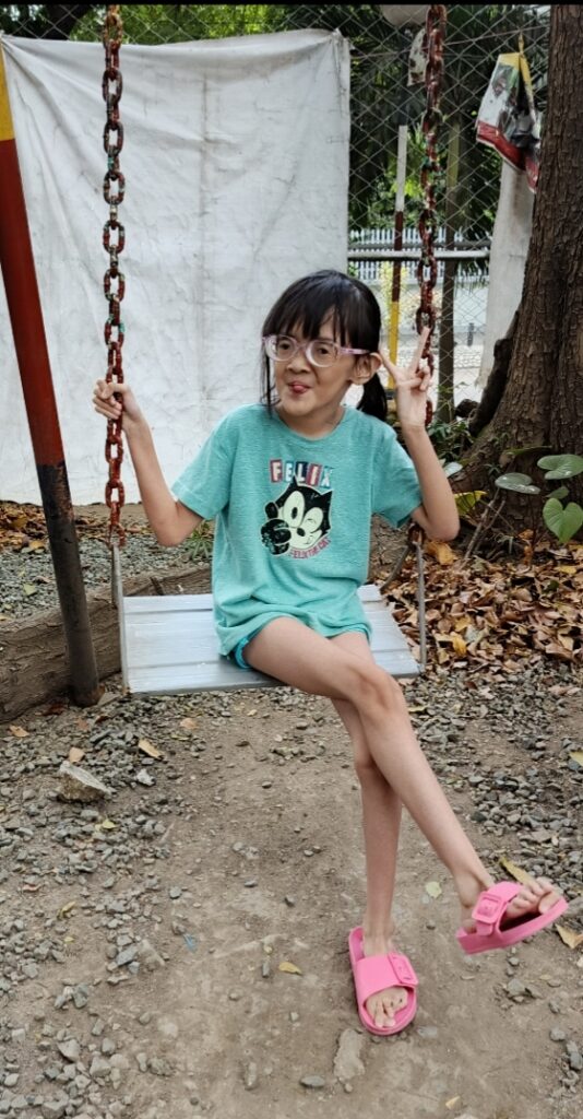 Destinee Jaye poses on a swing with pink Crocs and a light green top. She is wearing glasses, appears to show classic Marfan long-limb features, and is smiling flashing the peace sign. 