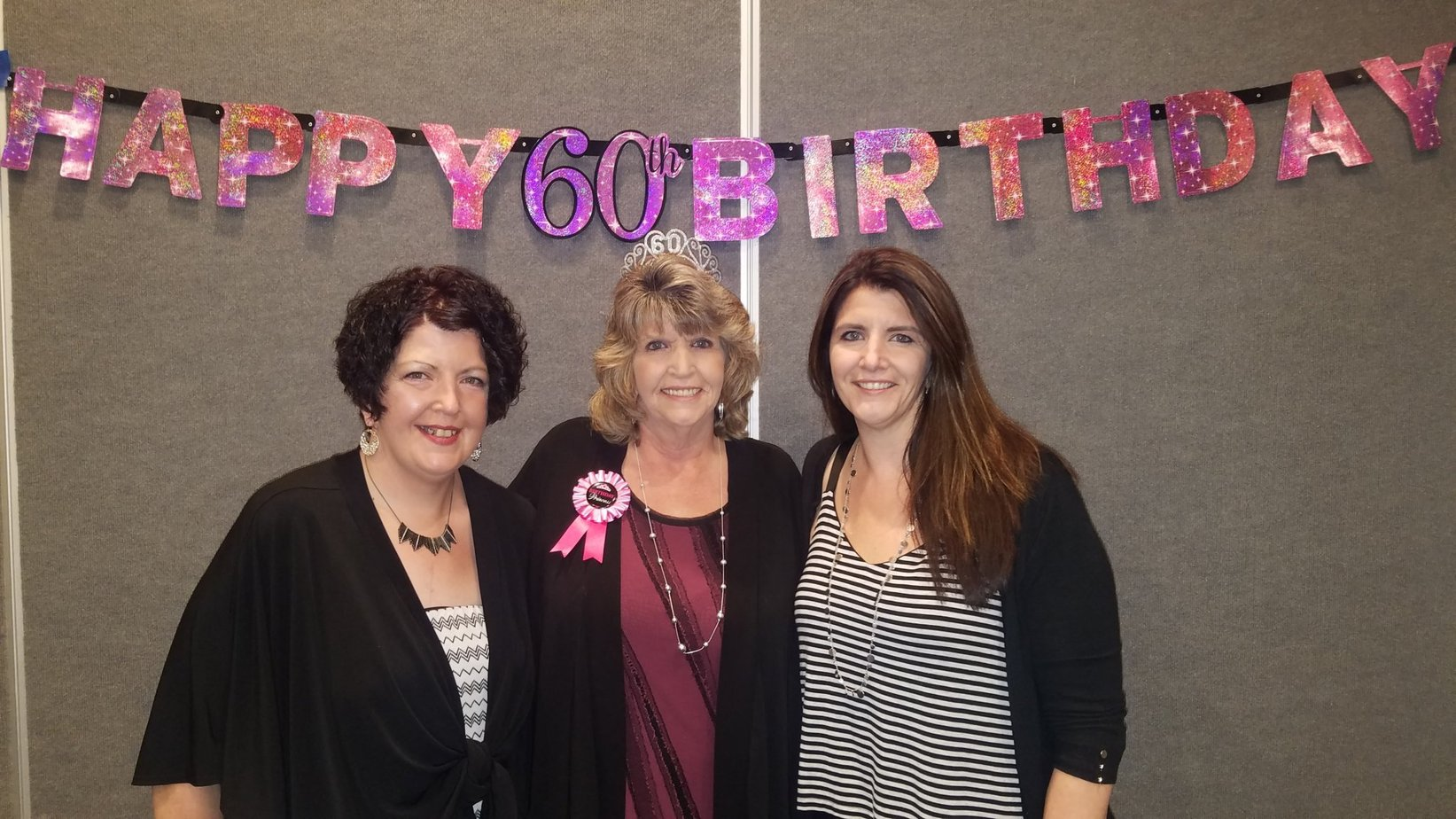 Steffy and Netty Mahnken and their mom celebrate her 60th birthday in front of a banner wearing celebratory clothes and mom in a crown.