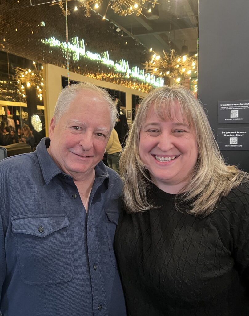 Tim Weil is pictured left in blue shirt with April Dawn Shinske right in black top. Behind them are the lights of the Broadway Museum as they discuss the life-saving potential of Marfan awareness through Tim's new book, Making Rent.