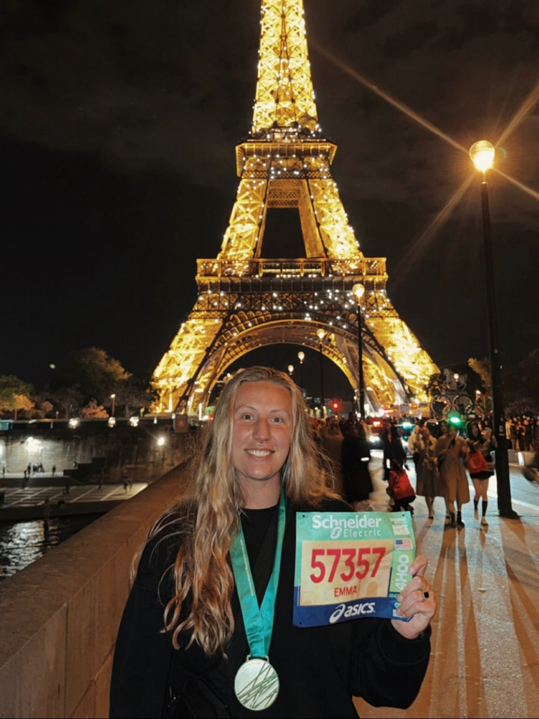 A runner is pictured standing in front of the Eiffel Tower at night with tower lit. She is wearing her medal and holding up her bib. 