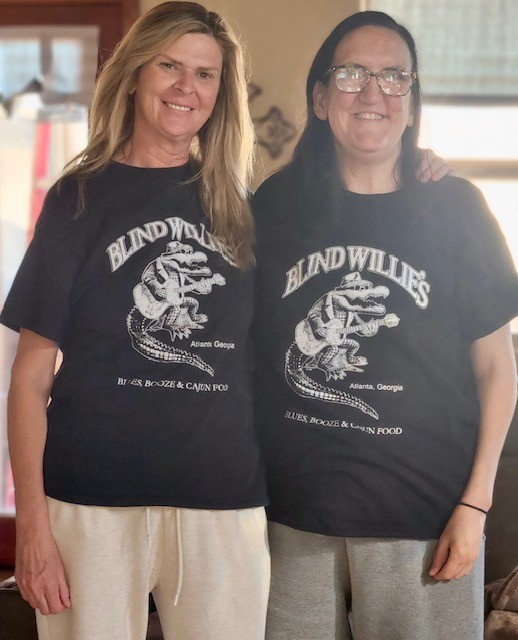 Danielle and Jennie, friends living with Marfan syndrome, pose smiling in matching black tee-shirts from a local eatery.