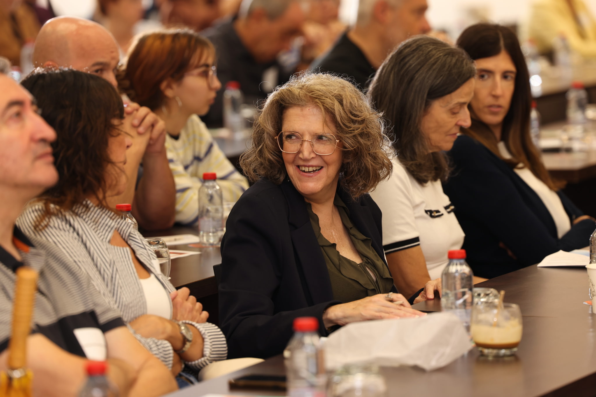 Asistentes sentados en mesas largas sonríen y escuchan durante el simposio de La Fundación Marfan en Barcelona.