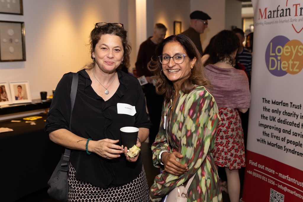 Two people stand smiling in front of a Marfan Trust sign at the 2026 London Symposium of the Marfan Foundation. 