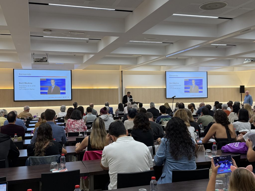 Una sala de conferencias llena de asistentes mira dos pantallas con una diapositiva de bienvenida para Bert Medina mientras habla desde el podio.