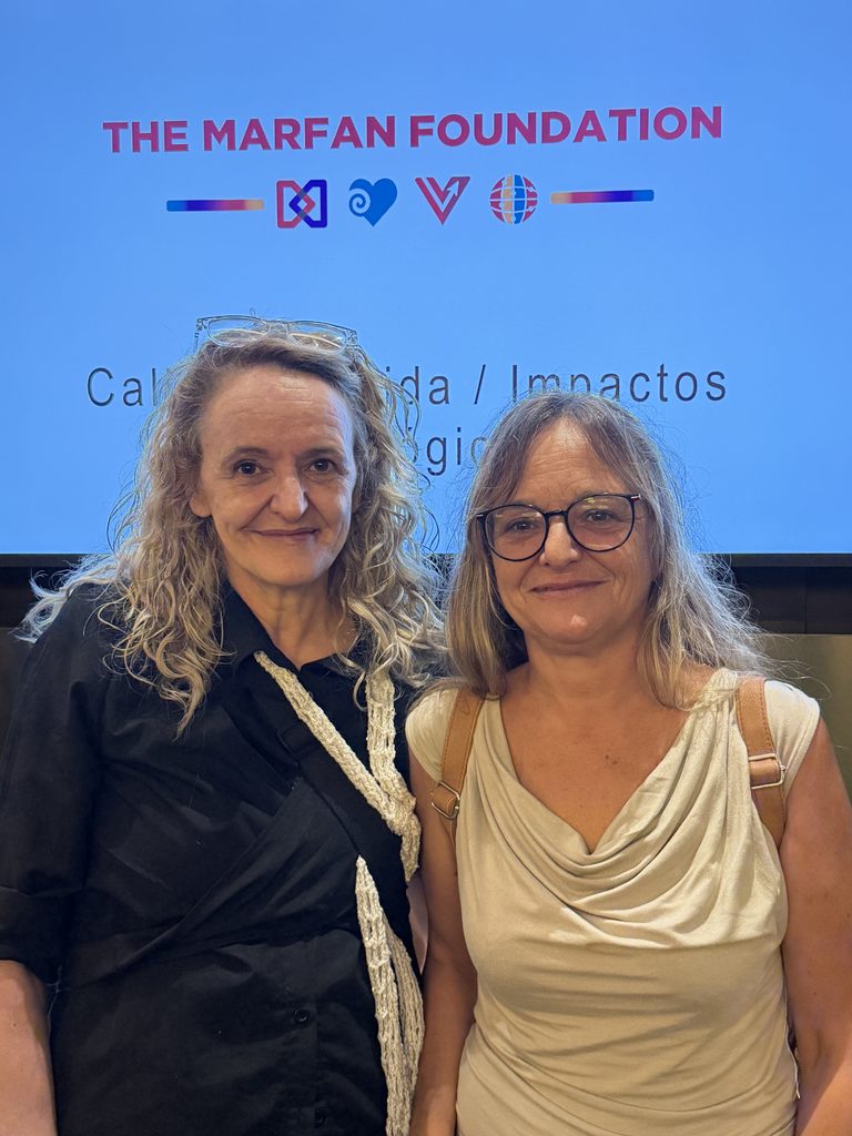 Mónica y su hermana Mercedes sonríen juntas delante de una pantalla con el logotipo de The Marfan Foundation durante el simposio en Barcelona.